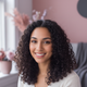Portrait of a smiling young adult woman with dark, curly hair and light brown skin, looking at the viewer. Blurred dusty rose